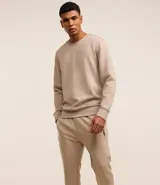 Conjunto deportivo color beige compuesto por un buzo de cuello redondo y un pantalón de jogging con cintura elástica y bolsillos laterales. El buzo es de corte clásico y el pantalón es ajustado en los tobillos.