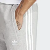 Pantalón de jogging Adidas Adicolor 3 Rayas, color gris con las tres rayas blancas a los costados. Cuenta con puños acanalados, bolsillos laterales con cierre, cintura elástica con cordón de ajuste y logo bordado.