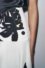 Falda midi de tiro alto confeccionada en hilatura de algodón color blanco con estampado floral en negro y amarillo. Tiene cierre con cremallera oculta en la costura y detalle de botones en la cintura.