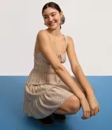 Vestido corto color beige, con escote recto, breteles medianos y lazo decorativo en el centro del busto. Presenta costura fruncida en la cintura y falda con babados en capas.