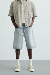 Bermuda de denim de algodón, color gris claro lavado, de corte ancho (baggy fit) y largo hasta la rodilla. Presenta cinco bolsillos y cierre frontal con cremallera y botón.