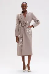 Abrigo largo color beige con corte recto y cintura anudada. Presenta motivos florales bordados a mano en tonos neutros y dorados en los bolsillos, mangas y bajo.