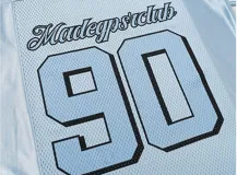 Remera estilo jersey de fútbol o americana, color celeste claro, con paneles de tela satinada y malla microperforada. Presenta cuello en V con ribete blanco, el número "90" grande en el frente con borde negro, y texto en la parte superior que dice "BAEN MADE" y "Madcapsclub".