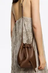 Bolso tipo bucket pequeño de piel vacuna, color marrón, con estructura blanda. Cuenta con asa regulable de piel y cierre de cordón ajustable. Presenta el logo metálico de BIMBA Y LOLA en dorado.