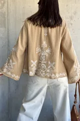 Chaqueta color beige con bordados blancos en el frente y las mangas, de corte recto y mangas anchas.