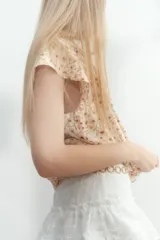 Blusa de tejido crepé color crema con estampado de flores pequeñas en tonos rosados y marrones. Tiene corte holgado, cuello con volados y escote en V con tiras para ajustar. Las mangas son cortas tipo mariposa con ribete de volado.