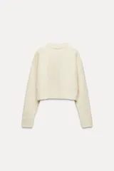 Jersey de punto color crema, de cuello redondo y manga larga. Bajo acabado con aberturas laterales.