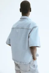 Camisa celeste de denim, corte amplio, con cuello clásico, manga corta, cierre frontal con botones metálicos, bolsillo de parche en el pecho y canesú en la espalda.