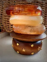Set de cinco brazaletes de diferentes materiales y colores: tres brazaletes rígidos de resina en tonos naranja y beige marmolado, un brazalete de madera marrón con apliques de estrellas y círculos dorados, y un brazalete de madera marrón liso.