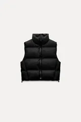 Chaleco negro acolchado con cuello alto y cierre frontal con cremallera. Tiene bolsillos delanteros con cierre de cremallera oculta y bajo ajustable con cordones elásticos laterales.