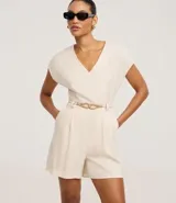 Enterito corto de mujer color beige claro, con escote en V, mangas cortas tipo capa, trabillas en la cintura y pliegues frontales. Incluye un cinturón fino trenzado de cuerda.