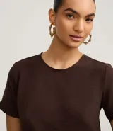 Blusa blanca de poliéster con cuello redondo, mangas cortas, cierre trasero en forma de gota, pequeñas aberturas laterales y acabados en pespuntes en las mangas y el cuello.