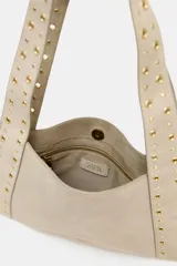 Bolso bandolera color beige de piel serraje, con tachuelas metálicas doradas en el cuerpo y las asas. Tiene doble asa de hombro, bolsillo interior y cierre con botón imantado.
