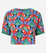 Remera de manga corta con cuello redondo y estampado repetitivo de loros rojos y hojas verdes sobre fondo azul y rojo. El borde de las mangas y el cuello es de punto acanalado verde.