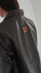 Blazer de cuero color gris oscuro, con aberturas laterales, aplique tipo pin desplegable bañado en bronce y botones de nácar.