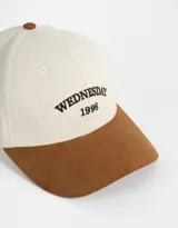 Gorra de béisbol bicolor, con cuerpo de canvas color crema y visera de gamuza color marrón. Tiene bordado frontal con la palabra "Wednesday" y el número "1996".