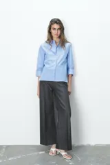 Camisa celeste a rayas verticales blancas, de cuello solapa y manga tres cuartos. Tiene aberturas laterales en el ruedo y cierre frontal con botones ocultos.