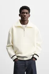 Sudadera color crudo de tejido técnico con cuello alto y cierre de cremallera. Tiene manga larga, corte relaxed fit, bolsillo tipo canguro y rib en puños y bajo con rayas negras.