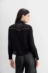 Camisa negra de manga larga, estilo texana, con cuello camisero y botones metálicos tipo snap. Presenta un bordado de tachas termo fijadas en forma de pico en la zona del pecho y hombros.