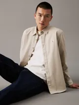 Camisa de manga larga color beige claro, con cuello clásico, cierre frontal con botones y logo de la marca bordado en el pecho.