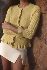 Chaqueta de punto amarilla con cuello redondo, manga larga y cierre frontal con botones. Detalle de volantes en los bordes.