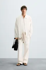 Camisa de corte relajado, color blanco hueso, con finas rayas verticales en tono grisáceo. Presenta cuello solapa, manga larga, bolsillo de parche en el pecho y bajo con aberturas laterales.