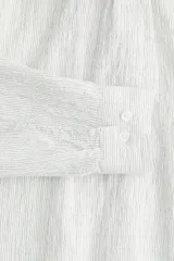 Camisa blanca de popelina de algodón texturizado, con cuello inglés, tapeta lisa, canesú con pliegue de caja en la espalda y mangas largas con abertura y cierre ajustable de botón en los puños. Corte holgado y bajo levemente redondeado.