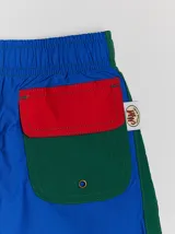 Short de baño azul con cintura elástica, rayas verdes en los laterales y bolsillo trasero con solapa roja y verde.
