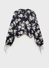 Blusa de satén color negro con estampado de flores blancas, cuello redondo, cierre frontal con botones, manga larga con puños elásticos y fruncidos, y corte recto.