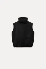 Chaleco negro acolchado, amplio, con cuello alto y sisa ancha. Tiene bolsillos laterales ocultos en la costura y cierre frontal cruzado con botones a presión.