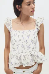 Blusa blanca con estampado floral en tonos lavanda, de tela ligera, con escote recto, breteles anchos con volados, espalda escotada con tiras finas horizontales que se atan en la parte superior, sección de punto smock en la espalda baja, costura fruncida en la cintura y peplum con vuelo.