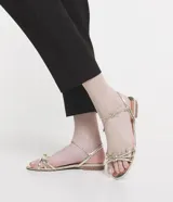 Sandalia plana marca Satinato, color dorado metalizado con textura tipo croco. Posee tiras finas con detalle de nudo en el empeine y cierre de lazo ajustable al tobillo. Diseño de puntera redonda y suela plana.