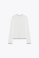 Chaqueta blanca de punto con cuello redondo y manga larga. Tiene vivos negros en el cuello, el borde frontal y los puños. Se cierra con botones en el frente.