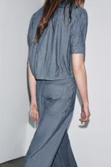 Camisa de denim azul con cuello redondo, manga corta abullonada con pliegues y bajo abullonado con pliegues. Cierre frontal con botones ocultos por solapa.