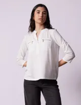 Blusa blanca marca Rio & Rian con cuello a la base y escote en v. Tiene dos bolsillos frontales con cierre metálico dorado y mangas tres cuartos.