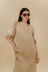 Vestido largo color beige, confeccionado en lino, con escote en V con volados y apliques de puntilla en las mangas. Posee bolsillos delanteros y tajos laterales.