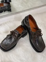 Zapato tipo mocasín de cuero color chocolate, con suela plana de goma negra, cordones al tono y costuras decorativas visibles en el borde superior.