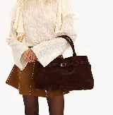 Cartera marrón de gamuza con doble asa corta, correa con hebilla decorativa y cierre superior.