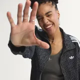 Campera deportiva Adidas Originals modelo Firebird, color negro con estampado animal print sutil. Presenta cuello camisero, cierre frontal y las icónicas tres tiras blancas en las mangas. Lleva el logo del trébol bordado en blanco en el pecho.