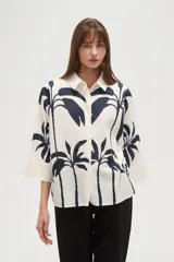 Camisa blanca con estampado de palmeras en color azul marino, de corte holgado, con cuello camisero, cierre frontal con botones y mangas 3/4 con puños anchos.