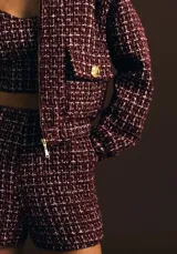 Chaqueta corta de tweed color borgoña con detalles en blanco. Presenta cuello redondo, cierre frontal con cremallera dorada, bolsillos delanteros y ribetes deshilachados.