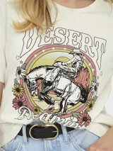 Remera color crema de algodón con cuello redondo y mangas cortas. Estampado frontal con un jinete a caballo, flores y texto "Desert Dreams Wild West".