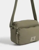 Bandolera gris claro de tela deportiva con bolsillo frontal, correa larga regulable, acceso con cierre superior y tag de goma.