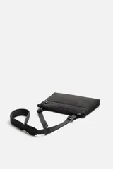 Bandolera negra fabricada en tejido técnico con correa ajustable. Tiene un compartimento principal con cierre de cremallera, un bolsillo interior con cremallera y un bolsillo frontal con cremallera y detalle de arnés.