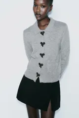 Chaqueta de punto gris con cuello solapa y manga larga. Tiene bolsillos delanteros de plastrón, rib en los acabados y cierre frontal con botones a presión en forma de lazo.