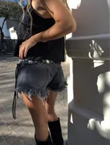 Short de jean negro con efecto acid wash, tiro bajo, con roturas y terminación deshilachada en el ruedo. Presenta detalles de ojales metálicos y tiras de ajuste laterales.