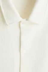 Camisa blanca de punto con textura acanalada, corte regular, cuello inglés, tapeta lisa, canesú en la espalda y bajo recto con aberturas laterales.