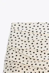 Falda pantalón corta color crudo con estampado de lunares negros, tiro medio, detalle de pliegues y volantes, y cierre lateral con cremallera.