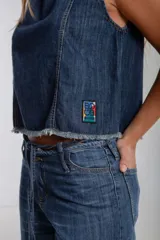 Blusa sin mangas de denim oscuro, con cuello redondo y costuras contrastantes en color marrón claro. Presenta un pequeño parche rectangular multicolor en la parte inferior frontal. El ruedo es deshilachado.