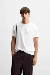 Camiseta blanca de corte holgado, cuello redondo y manga corta. Estampado frontal con texto en color negro y parche en la espalda. Detalle de pliegues en la espalda.